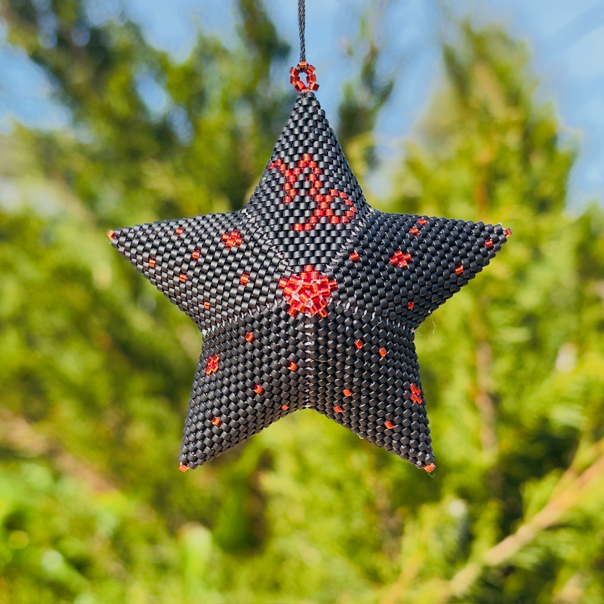 Beaded star ornament in dark charcoal with scattered red accent beads hanging outdoors